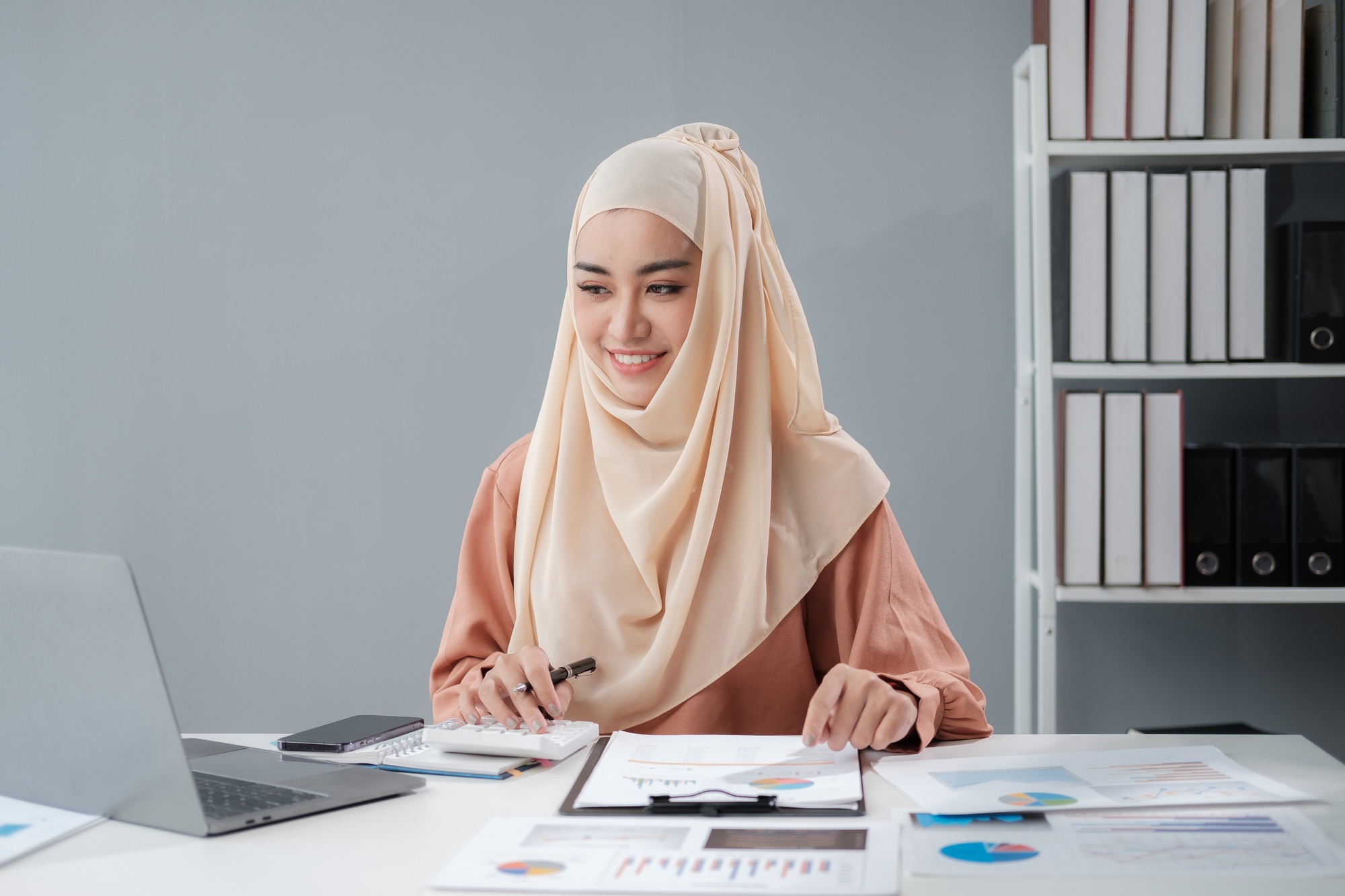 Muslim female economist working online with accounting documents on modern laptop device.